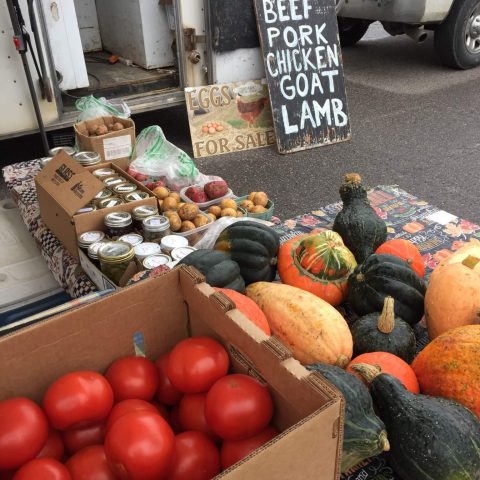 Stand with tomatoes and a variety of squash. Sign in the background lists beef, pork , chicken, goat and lamb.