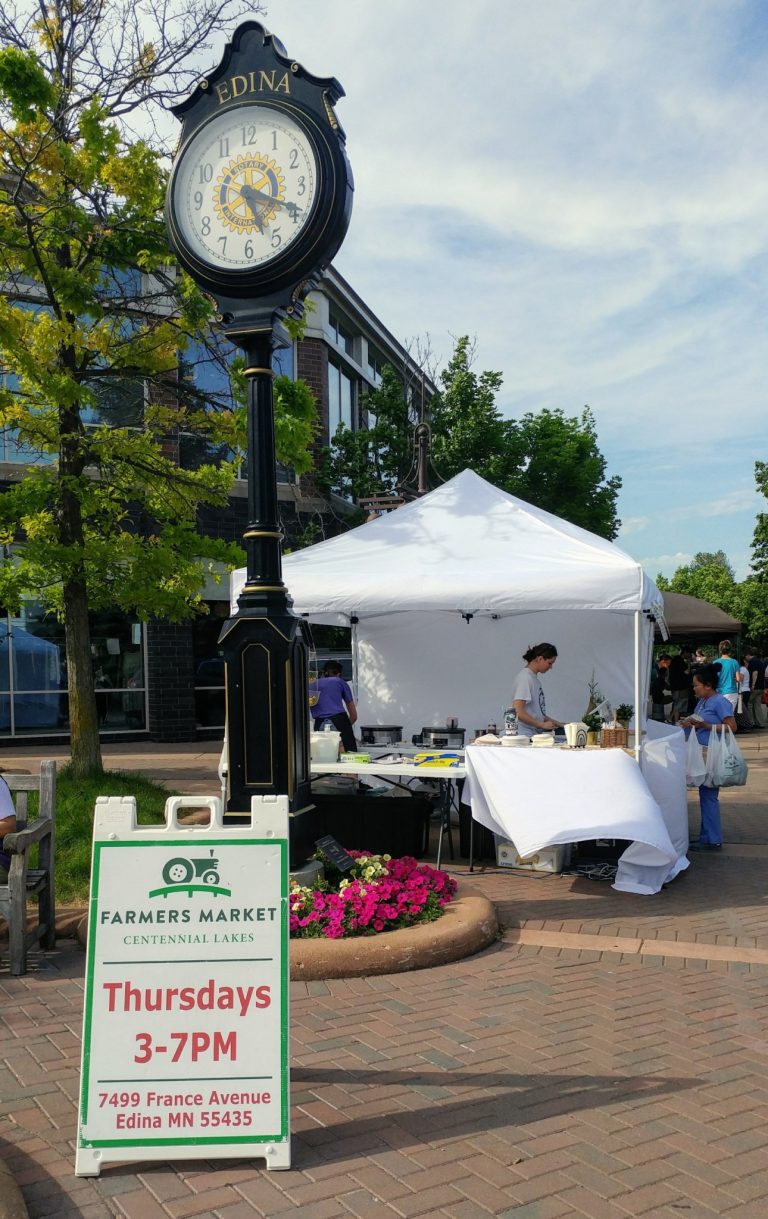 brick plaza with a farmer's market sign and tent. A clock tower stands tall at the left. Trees are in the background. Partly cloudy blue sky overhead.