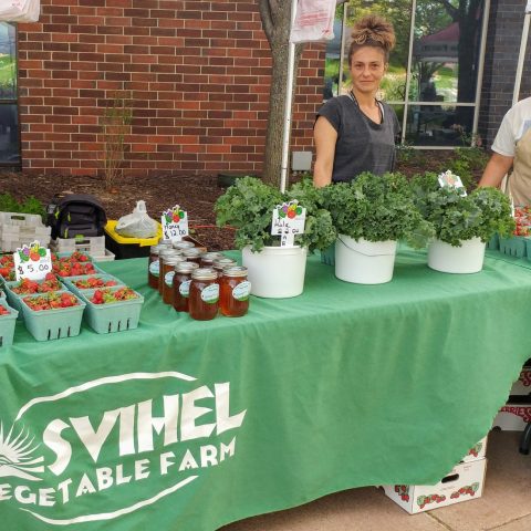Two women stand behind a green table with Svihel vegetable farm written on the front. strawberries, kale, and honey sit on the table. A brick wall is in the back with reflective windows. A white box peeks out through a slit in the green table cloth.
