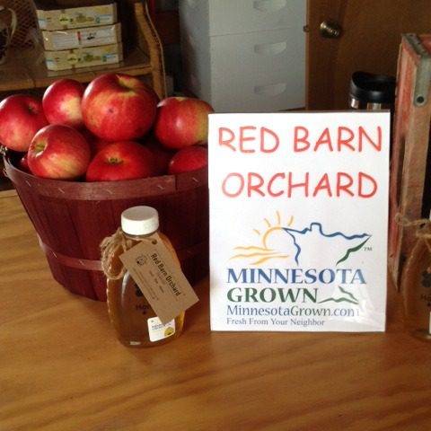 red barn and Minnesota Grown sign sitting next to a bottle of honey and a basket of apples