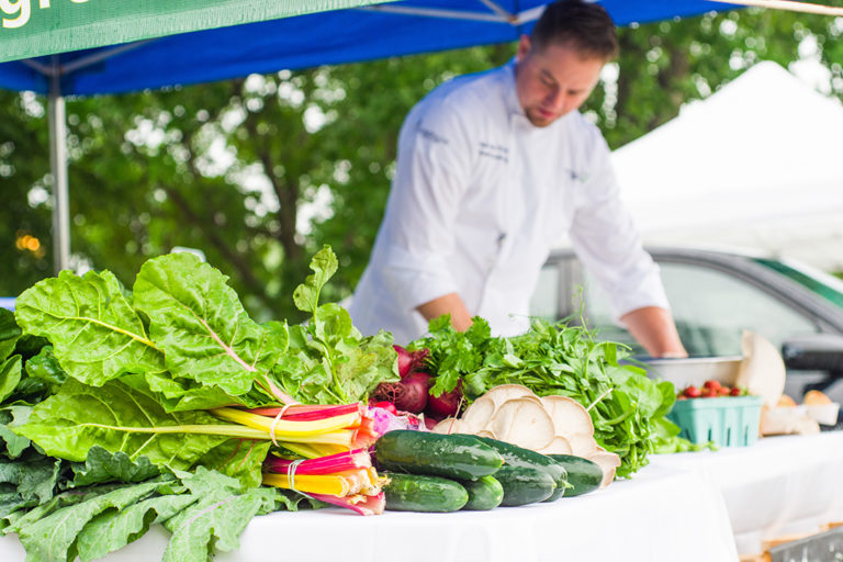 chef in the background prepping food and fresh produce such as cucumbers swiss chard and mushrooms in the foreground