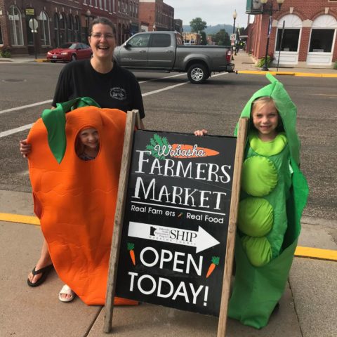Two kids dressed in peas and carrots costumes with the Wabasha farmers market sign