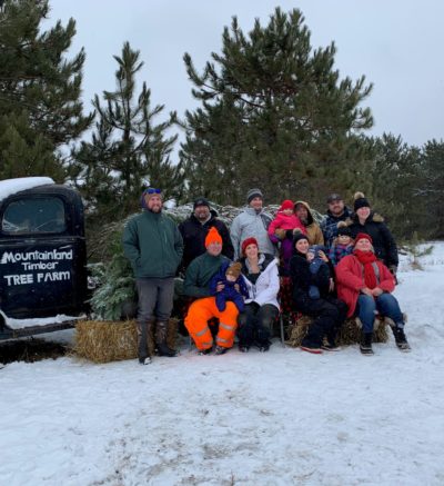 10 person family gathered for photo at the tree farm . the ground is covered in snow with pine trees in background