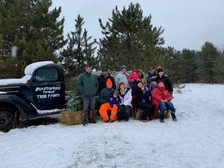 10 person family gathered for photo at the tree farm . the ground is covered in snow with pine trees in background