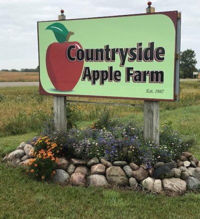 A large sign that reads "Countryside Apple Farm" and an image of a red apple with green stem. The sign stands in a field with landscaped plants, flowers, and rocks at its feet