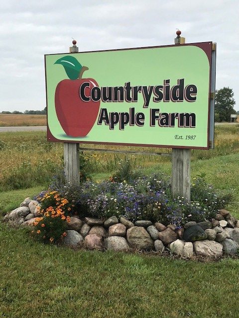 A large sign that reads "Countryside Apple Farm" and an image of a red apple with green stem. The sign stands in a field with landscaped plants, flowers, and rocks at its feet