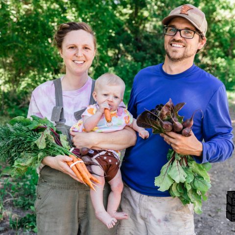 Smiling couple holding baby, carrots, and beets. Baby is chewing on a carrot