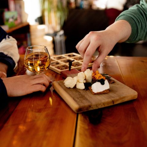 Close up of hands holding a glass of wine and picking cheese from a wooden board.