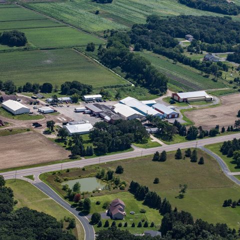 Aerial view of their farm. It features forests of trees, white buildings, and paved roads.