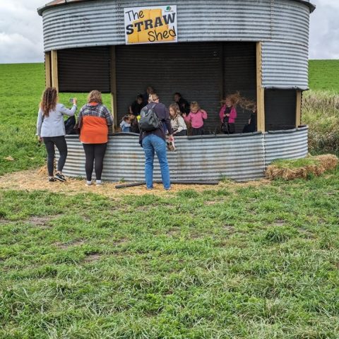 A metal shed labeled "Straw Shed" outside in the grass. People are standing outside of the shed talking to people inside the shed.