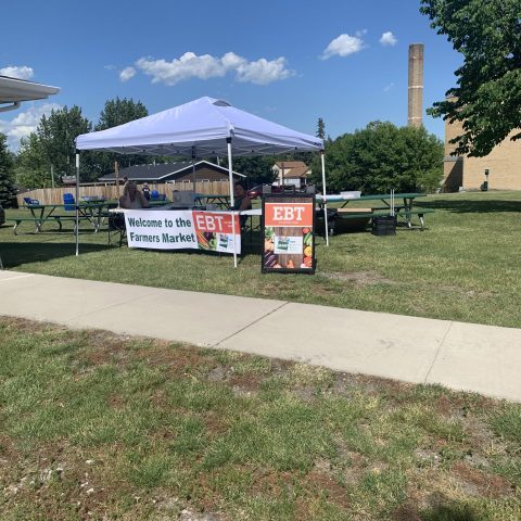 Picture of a farmers' market tent that has a banner in front of it. The banner says "Welcome to the Farmers Market EBT", implying that EBT is accepted at this market.