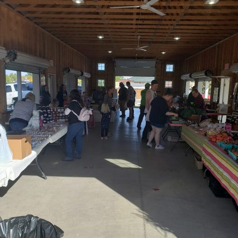 Picture of several farmers market stands indoors with people standing in front of them.