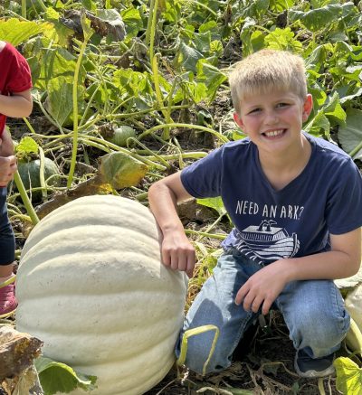 Young boy smiling beside a large, white pumpkin or gourd outside.