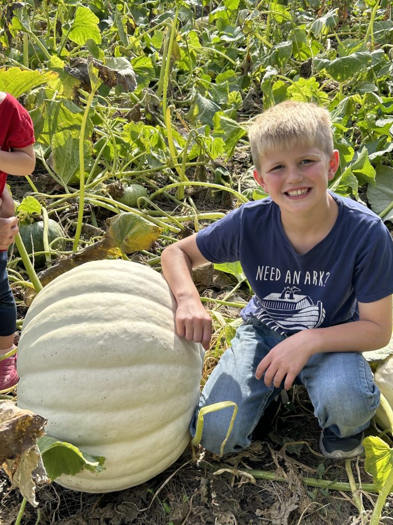 Young boy smiling beside a large, white pumpkin or gourd outside.