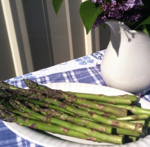 A plate of asparagus sitting on a blue tablecloth with a pitcher of lilacs near it.