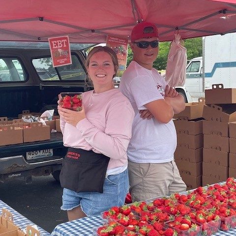 Two people stand back to back at a farmers market booth selling strawberries.