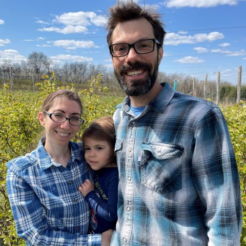 Picture of a smiling woman holding a young child and a smiling man beside them in a field outside.