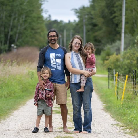 a young family smiles and poses, the wife holds a young child, the second child is at the husbands feet