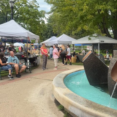 many farmers market tents and people milling about, there is a fountain in the foreground