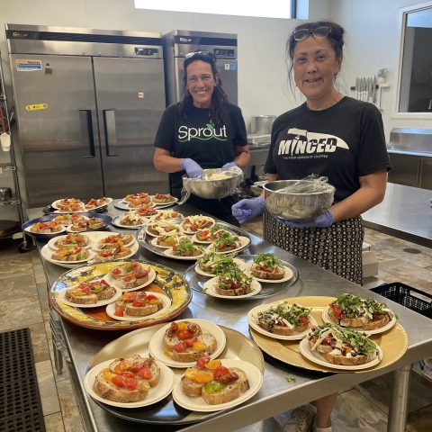 two women in a commercial kitchen smile as they garnish a table full of artisan toasts