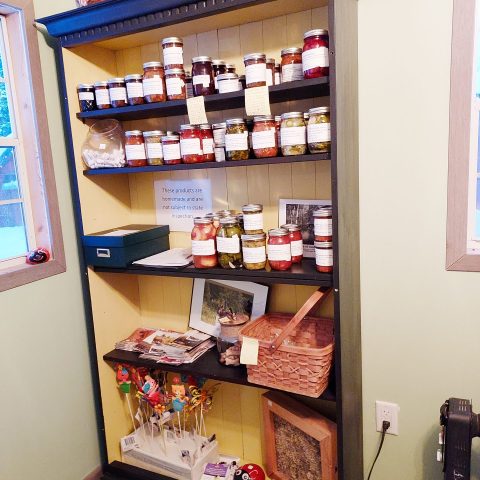 Shelving unit stocked with jarred goods, cash box and other items for sale.