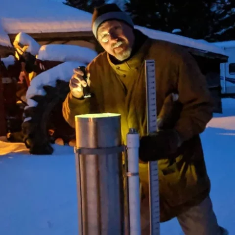 A man in the snow using a flashlight to take readings from a weather station.