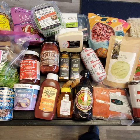 Picture of a basket of miscellaneous products (mostly food), including an array of condiments, cheese, salmon, a box of microgreens, fuzzy socks, and many more.
