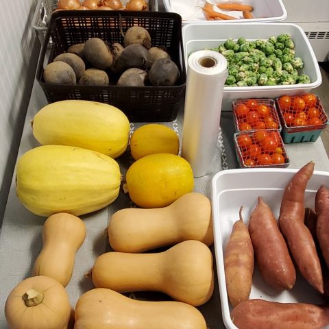 Picture of a table with fresh produce, including pumpkin squash, yellow melons, tomatoes, brussel sprouts, sweet potatoes, onions, and more.