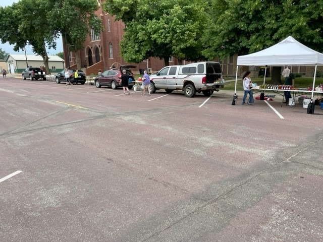 A picture of a parking lot with a farmers market vendor stand beneath a white canopy. There are people standing and cars parked around the stand.