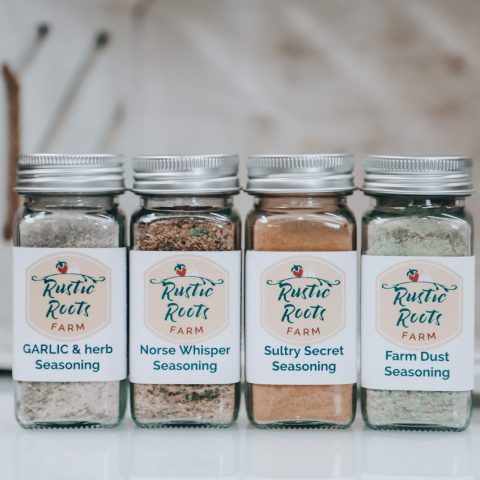 A row of four seasonings sit on a white platform against a white platform. The bottles say "Garlic & Herb Seasoning", "Norse Whisper Seasoning", "Sultry Secret Seasoning," and "Farm Dust Seasoning"