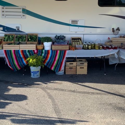 market tables with a bright tablecloth and filled with crates of fresh vegetables, canned goods, and baked items