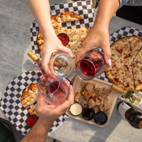 Three hands holding wine glasses toast over pizzas.