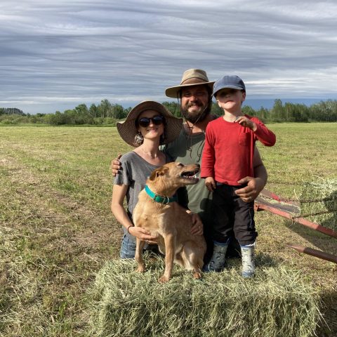 Family standing in a field. A small dog and a boy stand on top of a hay bale.