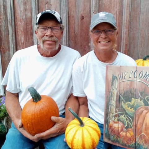 Picture of a man and a woman smiling in baseball hats and holding pumpkins and gourds.