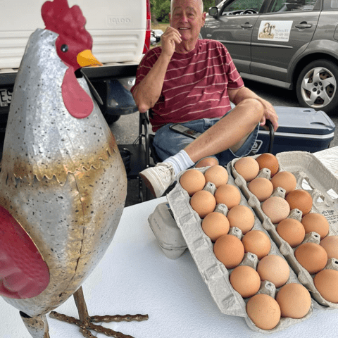 Picture of an elderly male smiling behind a farmers market table. On the table is 2 dozen brown eggs and a small metal chicken statue.