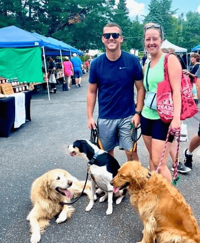 Picture of two farmers market goers smiling in the streets of the market with three dogs on leashes around them.