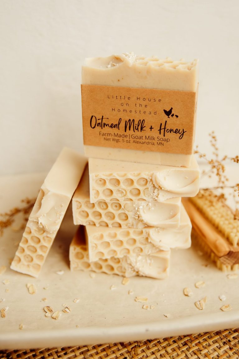 Beige bars of oatmilk and honey goatmilk soap are stacked on a beige surface. The bars have a honeycomb pattern in the top and a paper label.