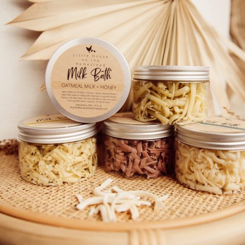 Clear jars of Oatmilk and honey milk bath body care products are stacked on a wicker table with a beige background