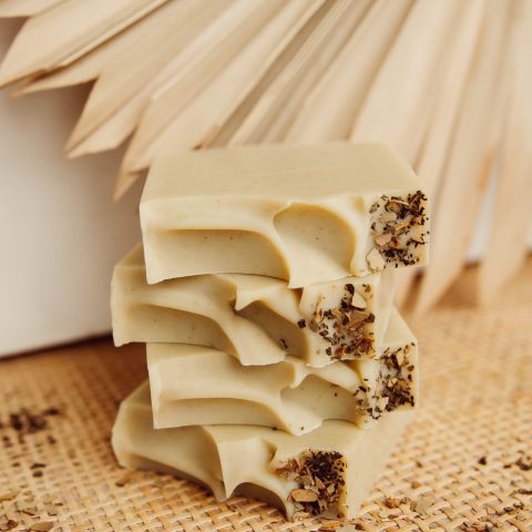 Stack of beige bars of soap on a rattan surface with folded paper adornment in the background, dried herbs adorn the soap and the background