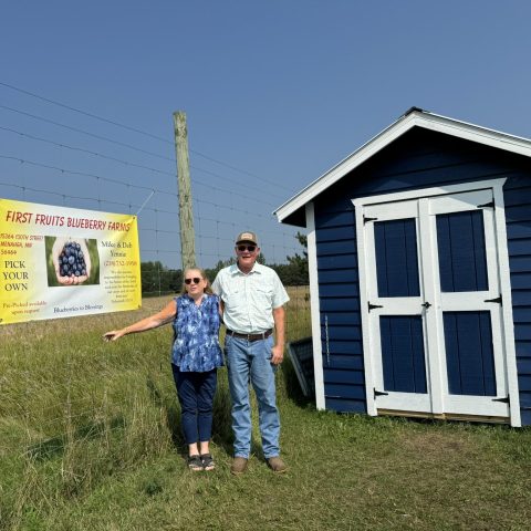 Deb and Mike Yennie posing for a photo in front of a bluew shed with white trim and a yellow poster for First Fruits Blueberry Farm