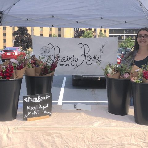 Picture of a woman standing at a Farmers' Market stand for Prairie Rose Floral and Farm. The stand features a table with bouquets of flowers in black vases. There is a banner with the Prairie Rose Floral and Farm logo behind the table.