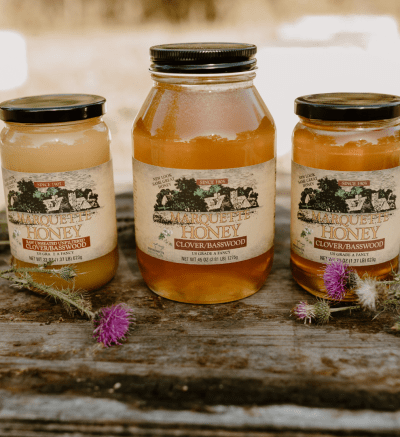 Three jars of honey on a wooden table. The table has small purple flowers on it. The honey in the jars vary in color from cream to golden, and the jars vary in sizes.