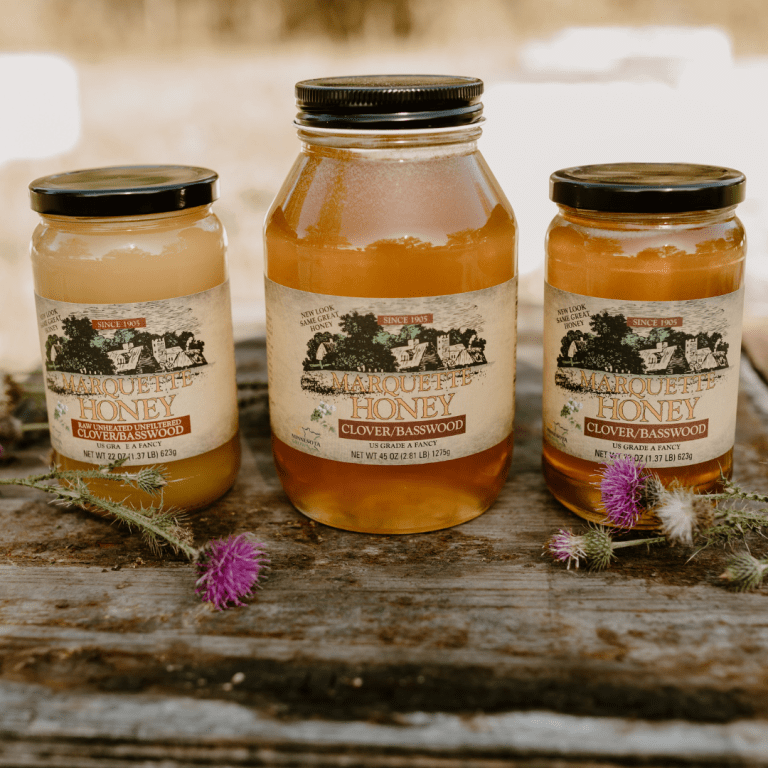 Three jars of honey on a wooden table. The table has small purple flowers on it. The honey in the jars vary in color from cream to golden, and the jars vary in sizes.