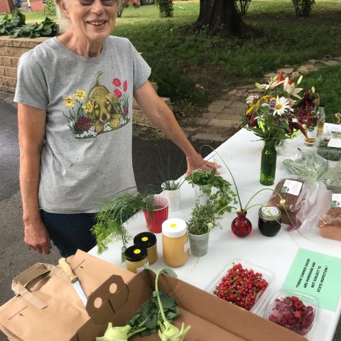 Picture of an elderly woman standing before a farm stand with flowers and fruit on the table.