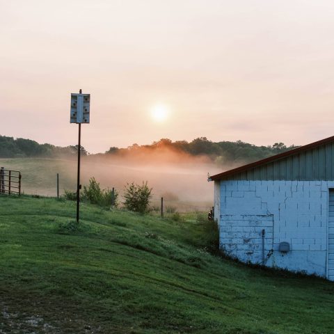 A sunrise with a farm building and a green hill covered in fog.