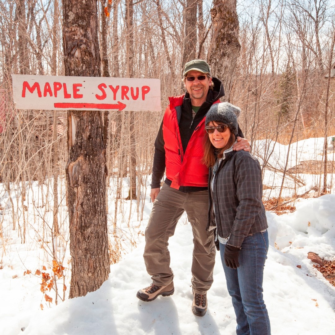Two people standing in a snowy forest next to a sign that says Maple Syrup