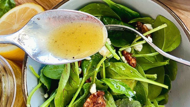 Close-up of a spoon with a yellow vinaigrette in it, hovering above a green salad with pecans.
