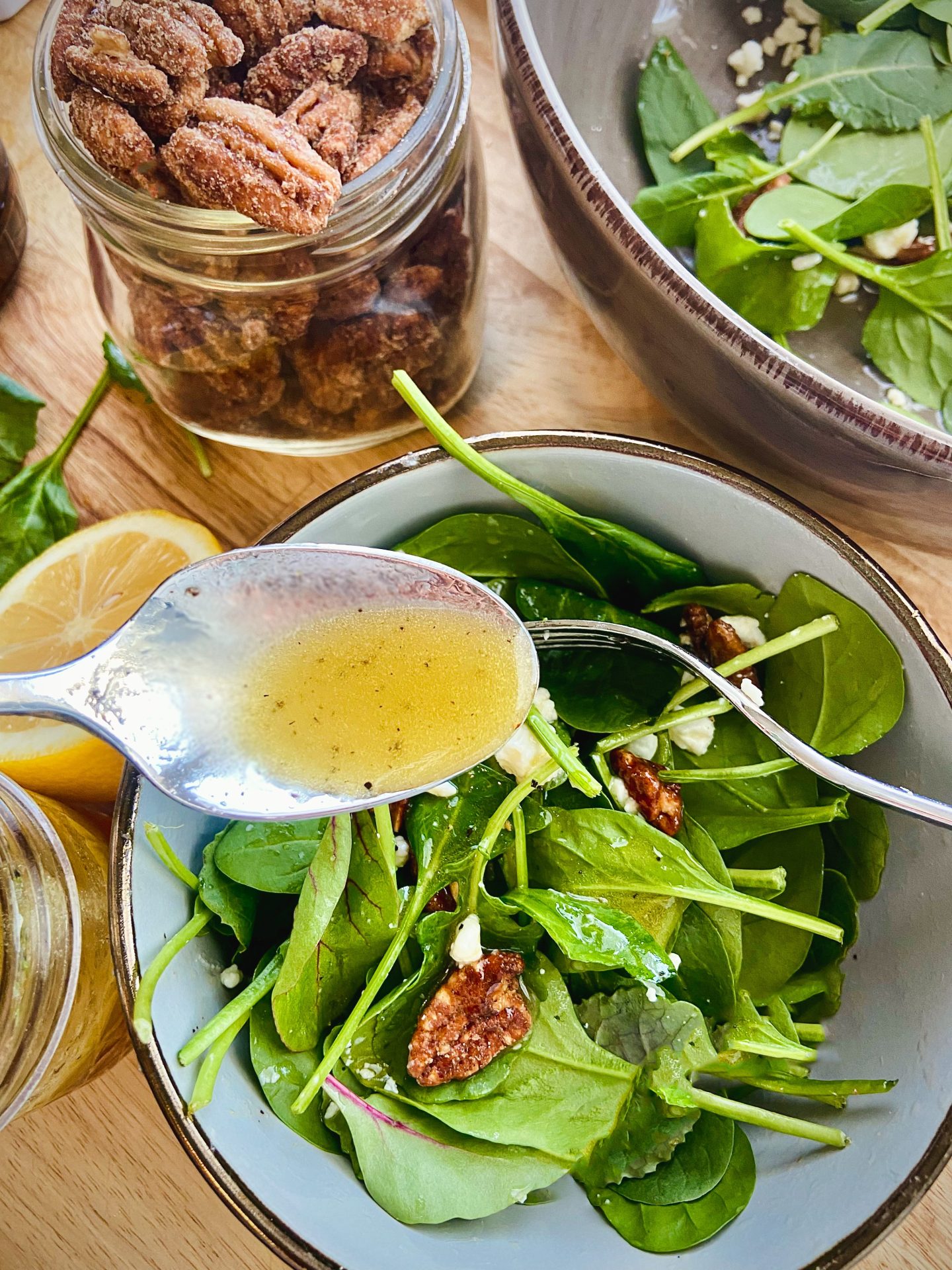 Overhead shot of a green salad with pecans and goat cheese. In the upper left corner is a jar of candied nuts and spoon filled with dressing hovers over the salad.