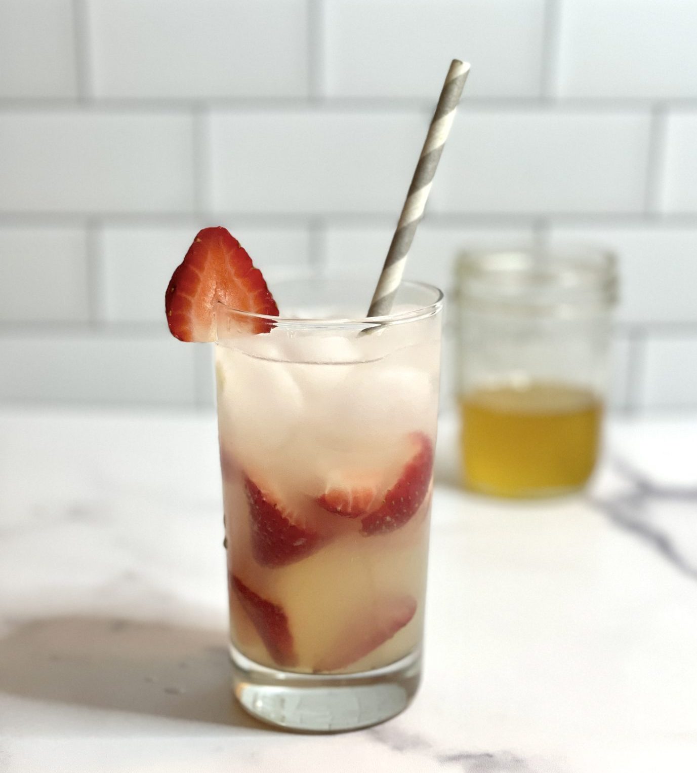 A highball glass with a strawberry filled cocktail. In the background is a jar of honey.