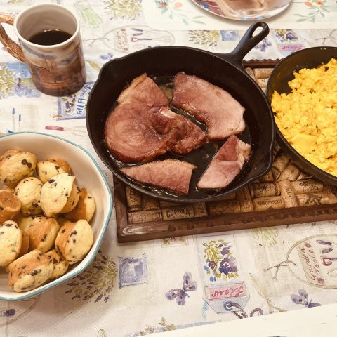 Picture of a homemade breakfast with chocolate chip muffins in a bowl, a cast iron pan with ham in it, and a pan of scrambled eggs.